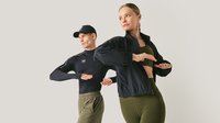 Zwei Personen in Sportbekleidung posieren mit gebeugten Armen vor einem schlichten, hellen Hintergrund, sie tragen schwarze Oberteile und olivgrüne Hosen.