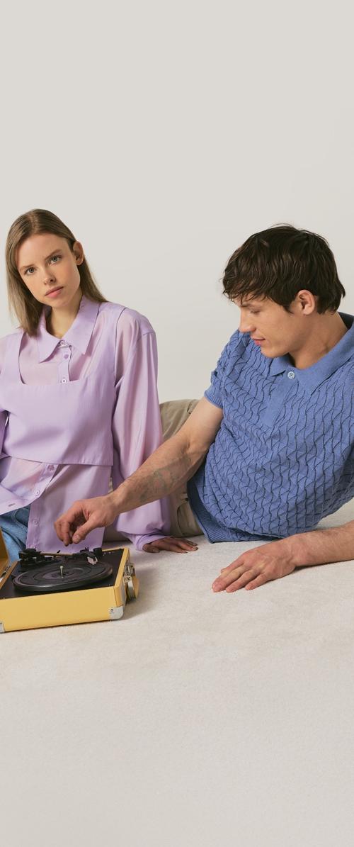 Jonge man die een gele draagbare platenspeler afstelt terwijl een vrouw in een lavendelblouse toekijkt, beiden zittend op een lichte vloer tegen een eenvoudige achtergrond.