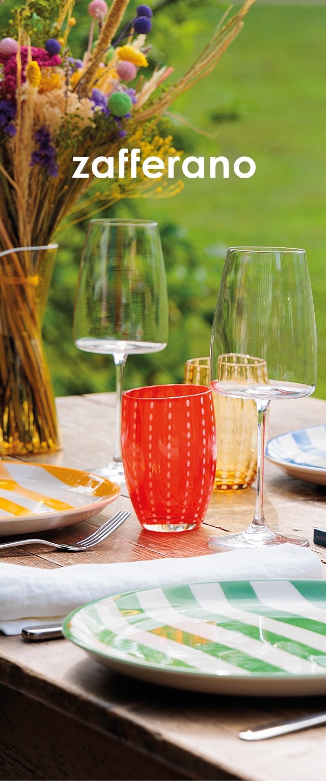 Colorful dinnerware set on a wooden table. Features striped green and white plates, textured glassware in red and amber, and a floral centerpiece.