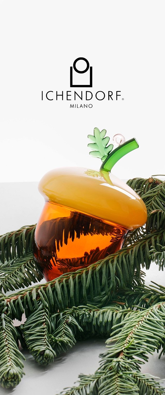 Pieza decorativa de vidrio en forma de bellota, en tonos ámbar y verde, con un cuerpo texturizado y un acento en forma de hoja. Presentada sobre ramas de pino.