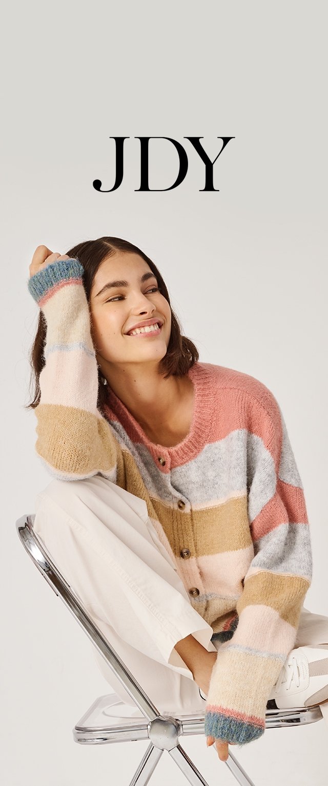 Junge Frau lächelt und trägt eine mehrfarbige, gestreifte Strickjacke, cremefarbene Hose und weiße Sneaker, während sie auf einem modernen Stuhl sitzt.