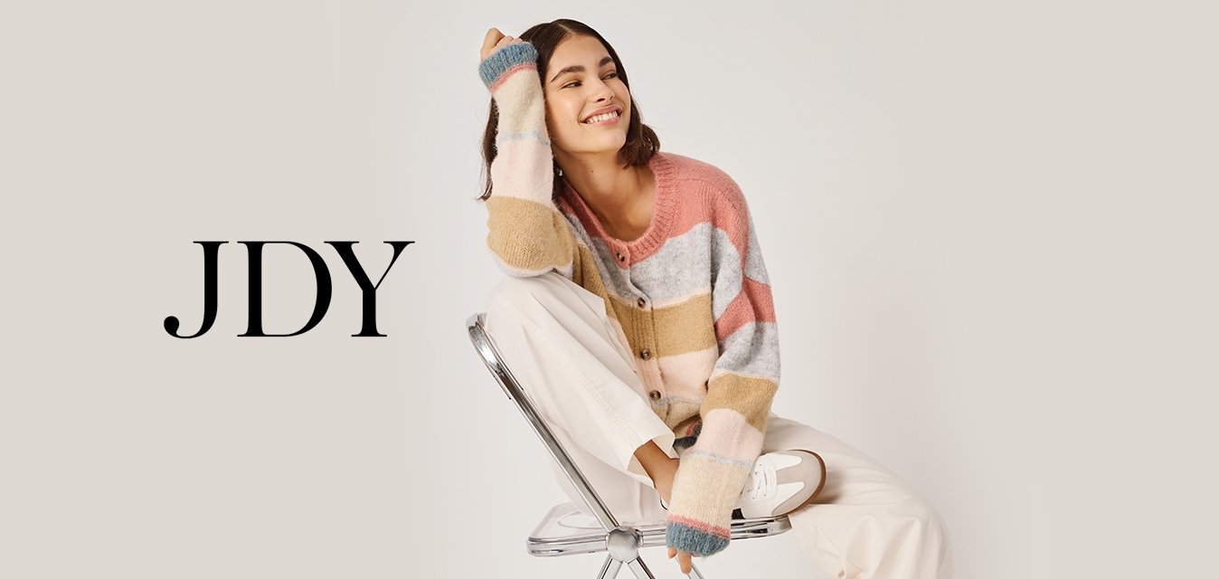 Mladá žena se usmívá, sedí bokem na kovové skládací židli, má na sobě vícebarevný pruhovaný svetr, bílé kalhoty a tenisky.