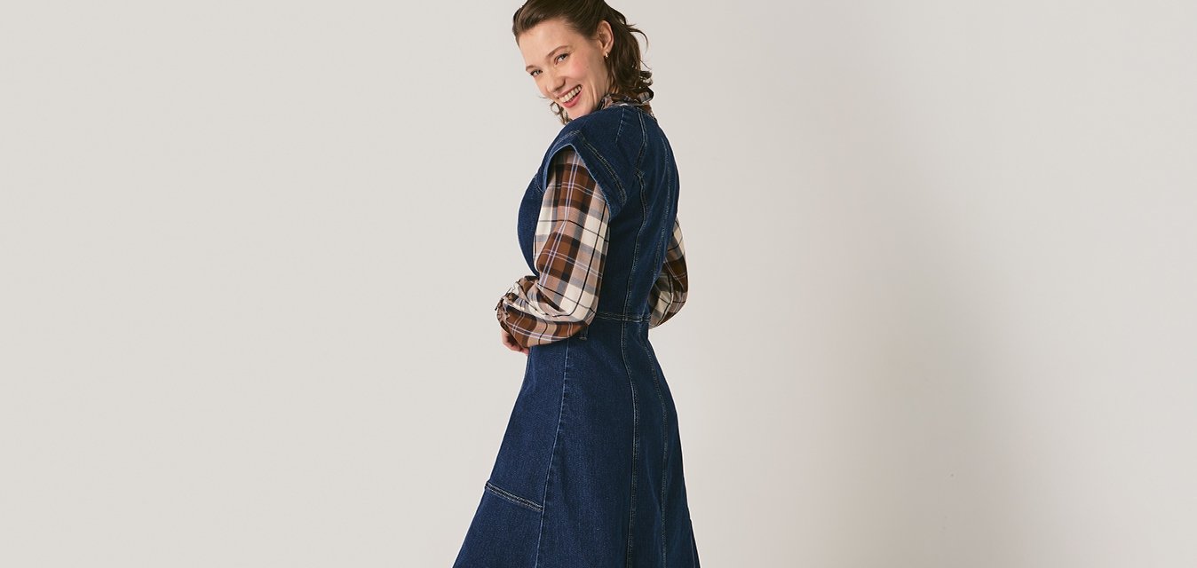 Žena v dlouhých tmavě modrých džínech nad košilí s kostkovaným vzorem, obuta v hnědých botách, stojící a usmívající se zpět na jasném světle pozadí.