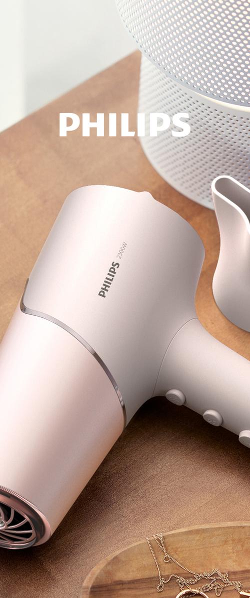 Baltas „Philips“ 2300W plaukų džiovintuvas, baltas perforuotas cilindro formos garsiakalbis, balta keramikinė vaza ir medinis padėklas su aukso papuošalais ant rudos paviršiaus.