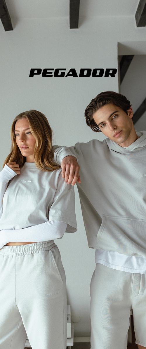 Jeune homme et femme en tenue de sport décontractée gris clair posant à l'intérieur sous le mot « PEGADOR » sur un mur blanc avec des poutres apparentes.