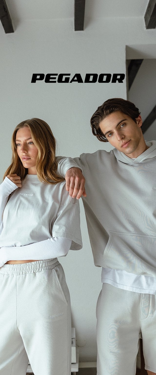 Joven hombre y mujer en ropa deportiva casual de color gris claro posando en interiores bajo la palabra "PEGADOR" contra una pared blanca con vigas expuestas.