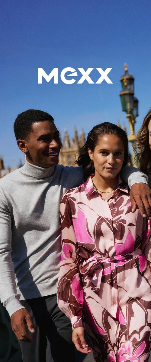 Ein Mann in einem grauen Rollkragenpullover mit dem Arm um eine Frau, die ein rosa-braunes Blumenkleid trägt, steht draußen unter klarem blauem Himmel.