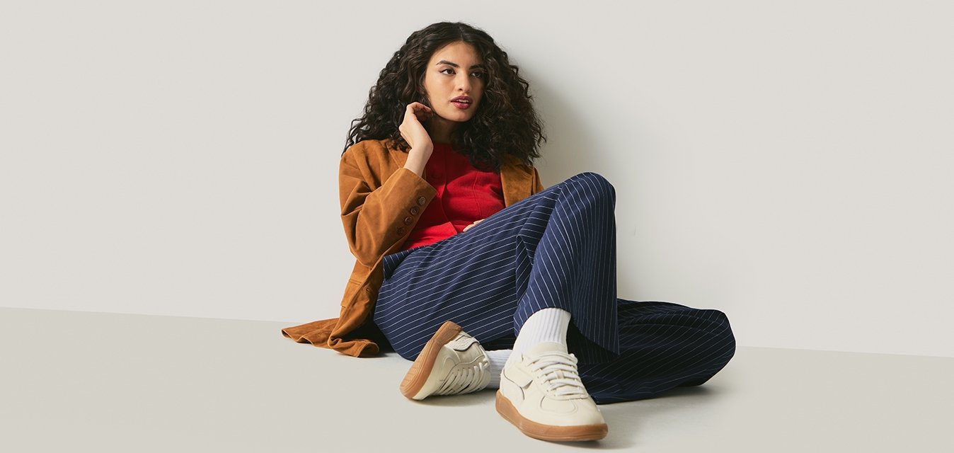 Mladá žena s kudrnatými vlasy, která má na sobě hnědou bundu, červené tričko, tmavě modré kalhoty s proužky a bílé tenisky, sedí na zemi opřená o bílou zeď.