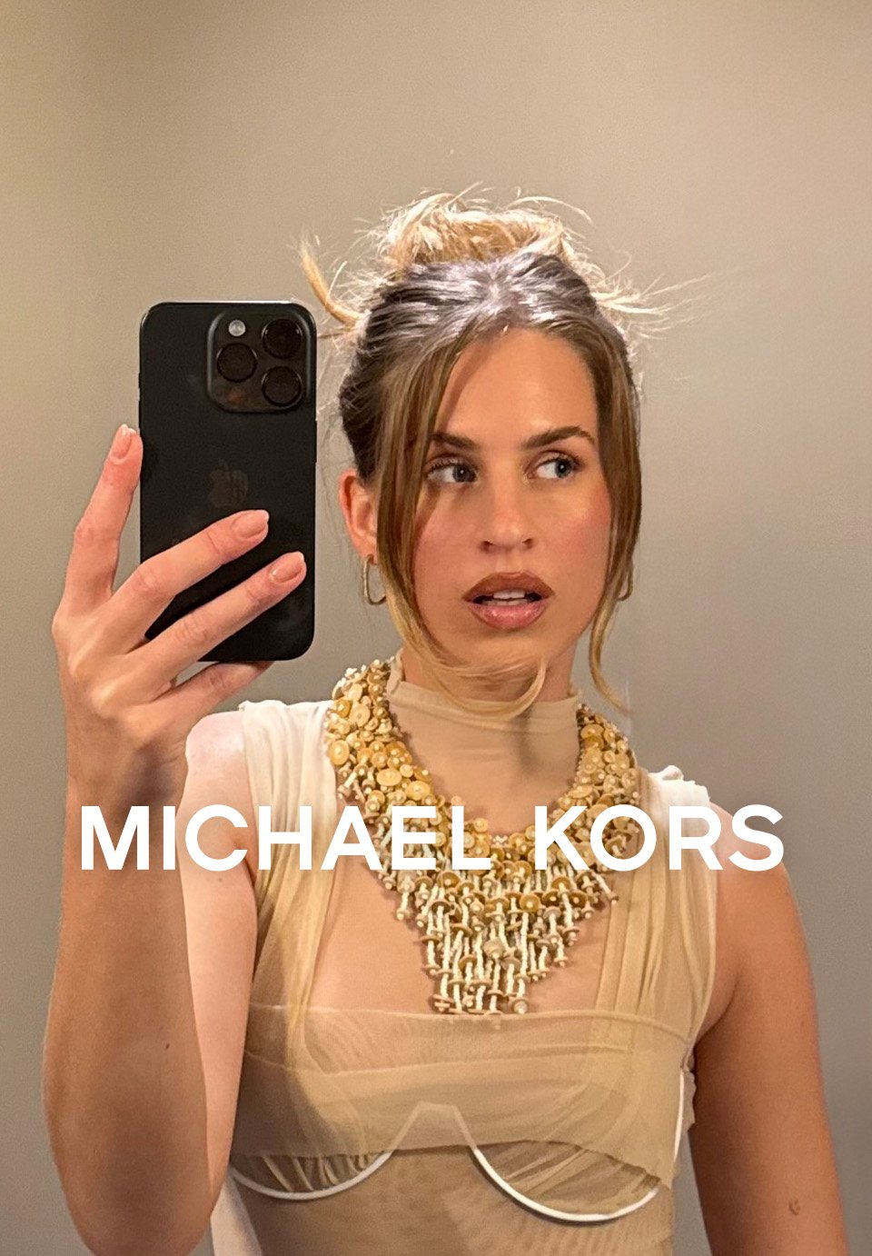Selfie miroir d'une femme en tenue beige Michael Kors et collier de coquillages, tenant un smartphone noir.