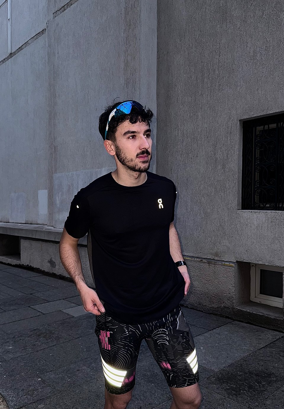 Bărbat în tricou de alergare negru și pantaloni scurți cu model, stând pe trotuar cu ochelari de soare pe cap.