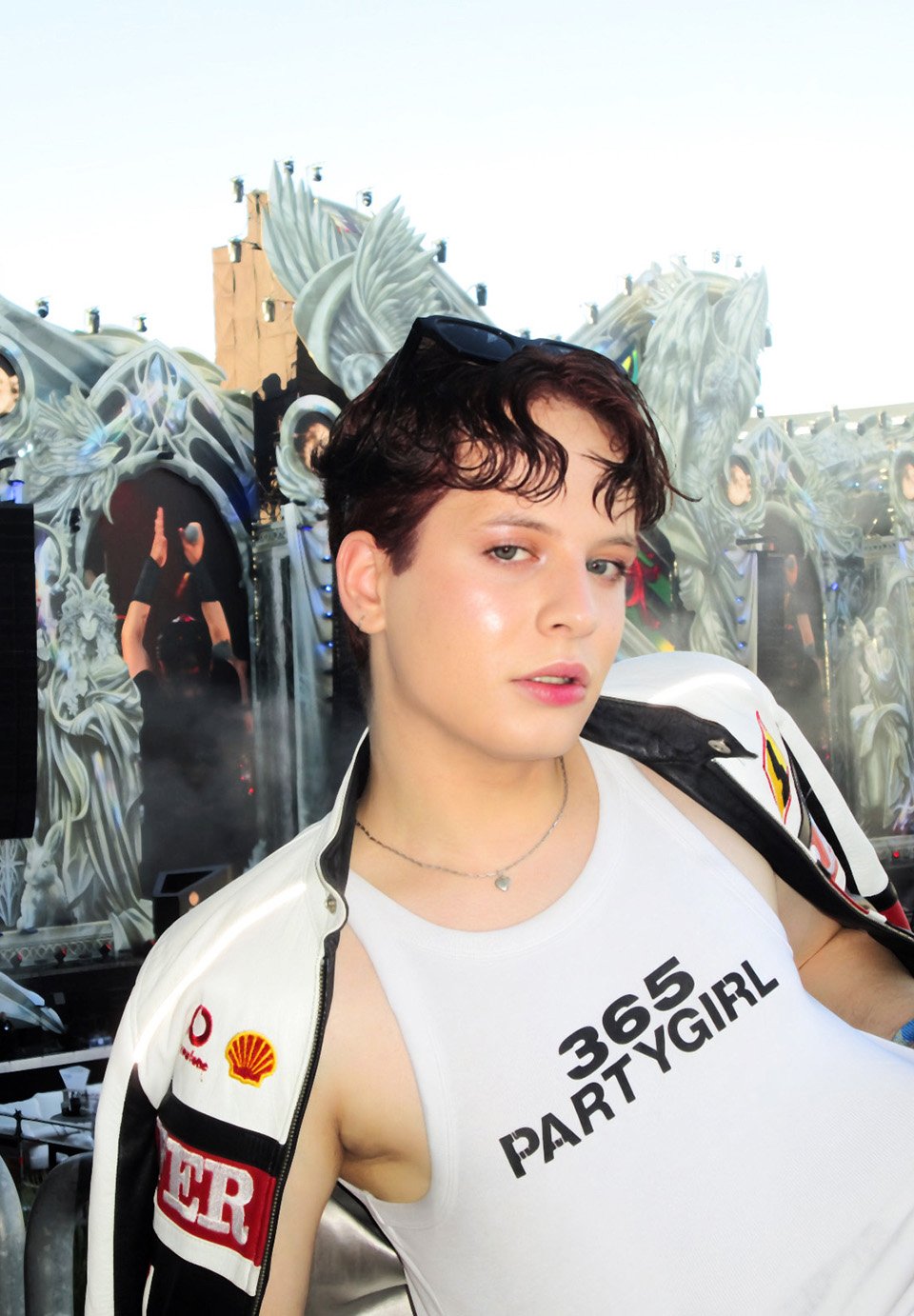 O persoană în maiou cu textul „365 PARTYGIRL” și jachetă de curse pozează în fața unei scene complexe de festival în aer liber.