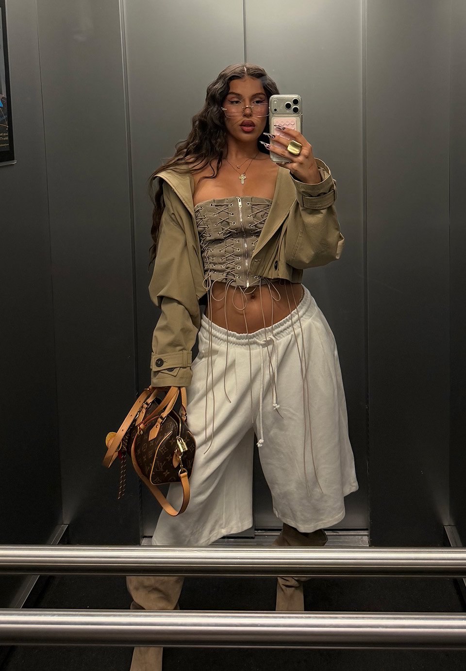 O femeie în corset kaki cu șireturi și pantaloni de trening albi își face un selfie în oglindă în lift, ținând o geantă de firmă.
